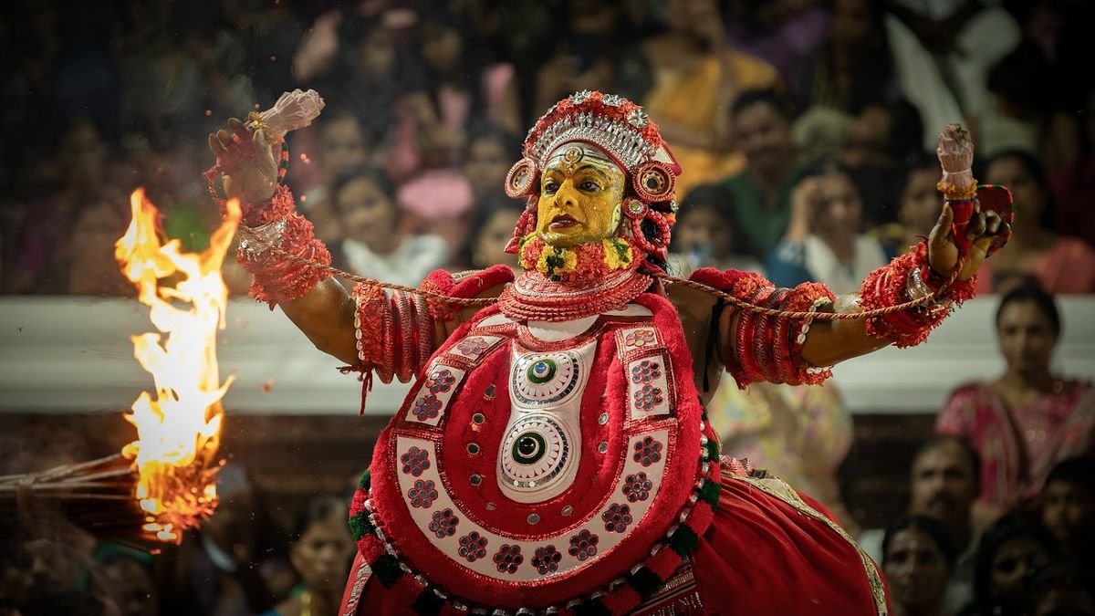 A Theyyam in red attire holding a flame, with devotees watching from behind
