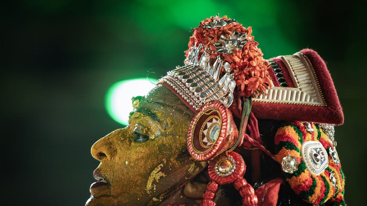 Profile of a Theyyam in a silver crown and red and green attire against a green background