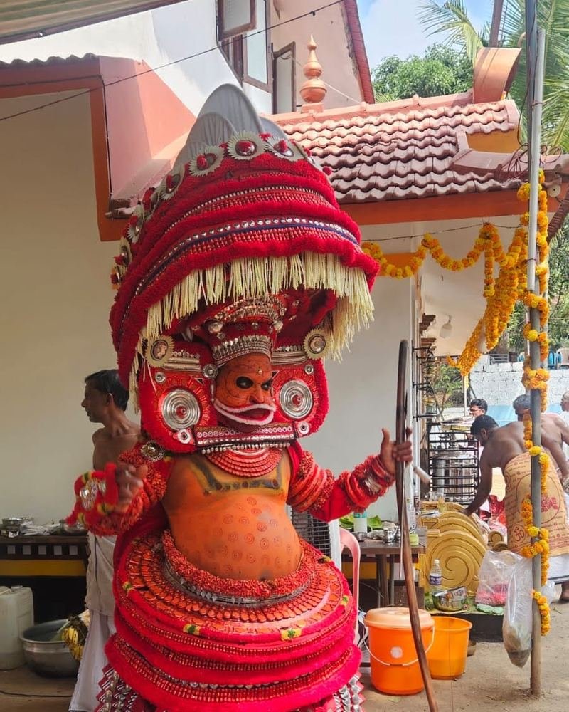 A Theyyam in a red cone headdress at a family shrine by day in Kannur
