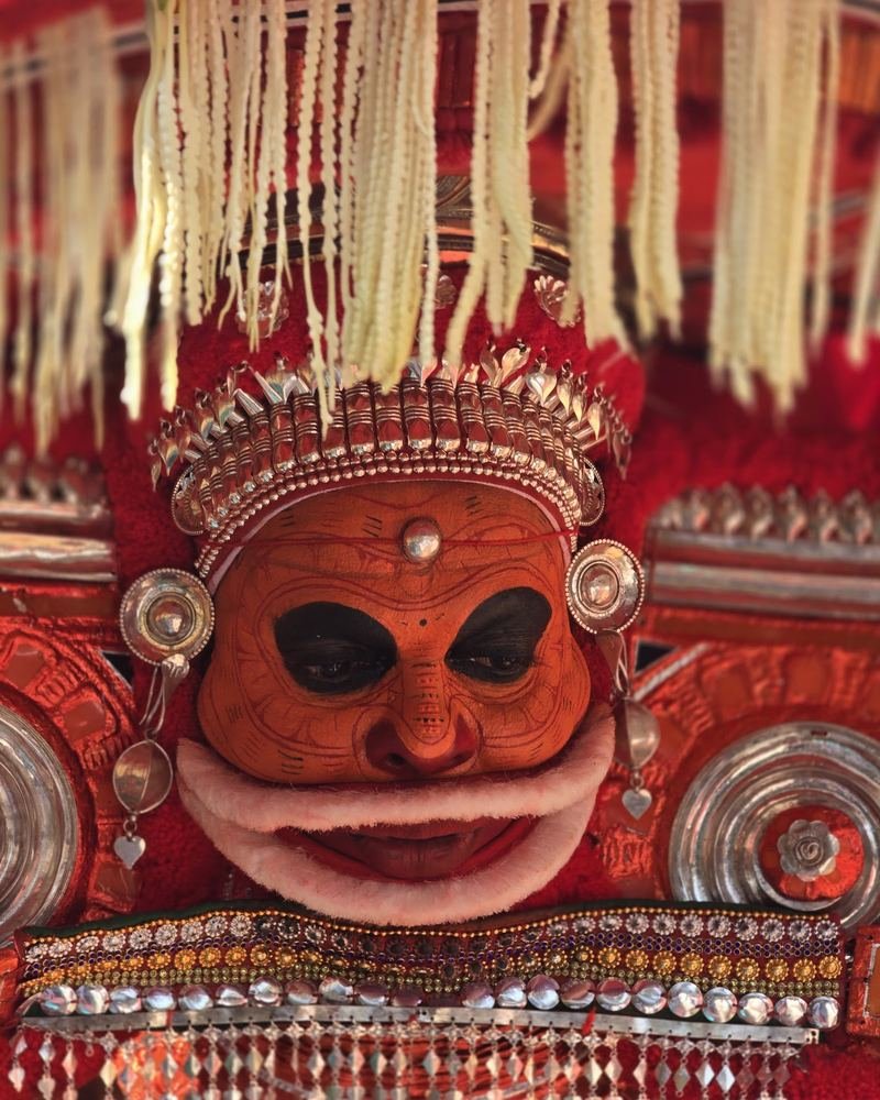 Close portrait of a Theyyam with orange face, silver ornaments and a white fringe