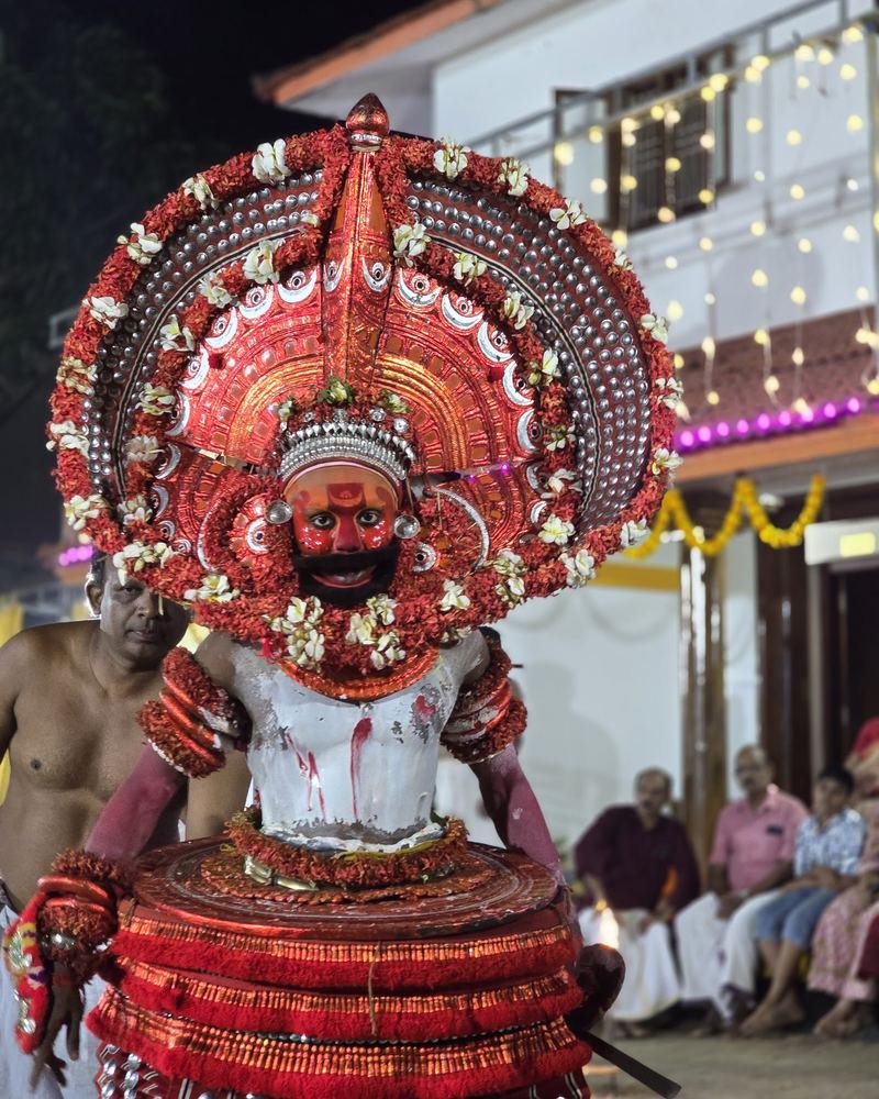 A Theyyam in a large red and silver headdress with flowers, at a shrine in Kannur