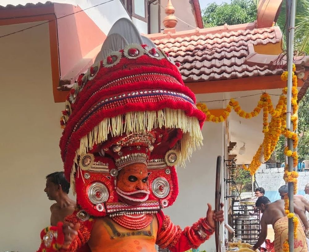 A Theyyam in a red cone headdress at a family shrine by day in Kannur