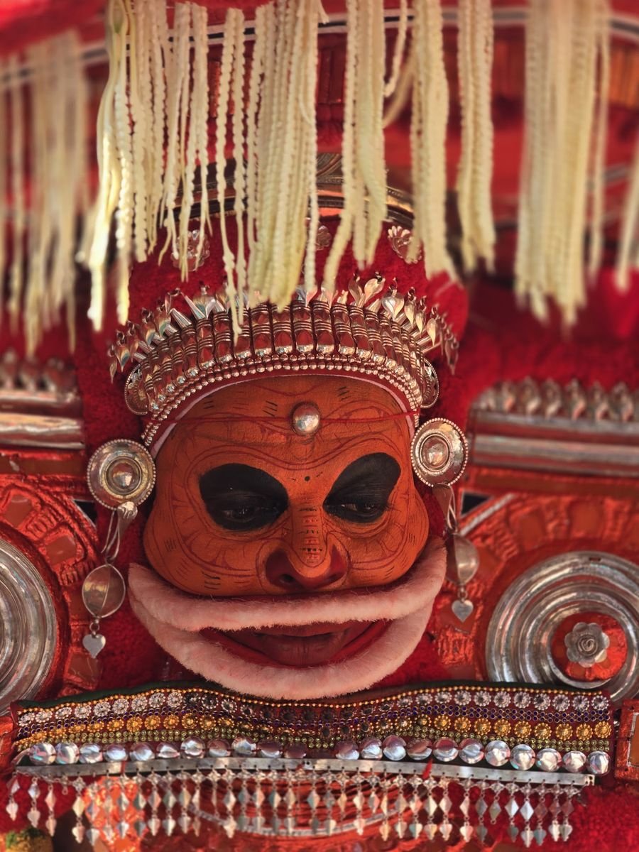 Close portrait of a Theyyam with orange face, silver ornaments and a white fringe