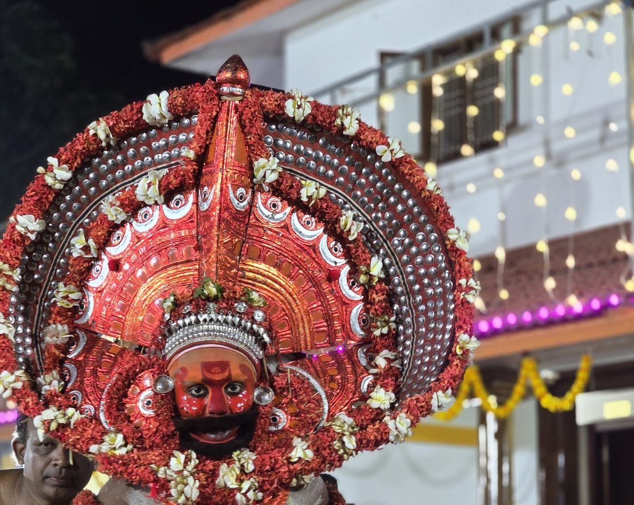 A Theyyam in a large red and silver headdress with flowers, at a shrine in Kannur