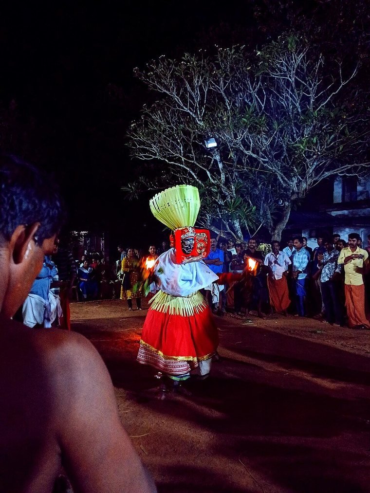 Karuval Bhagavathi Theyyam in red and white attire with a green headdress, at night in a shrine yard