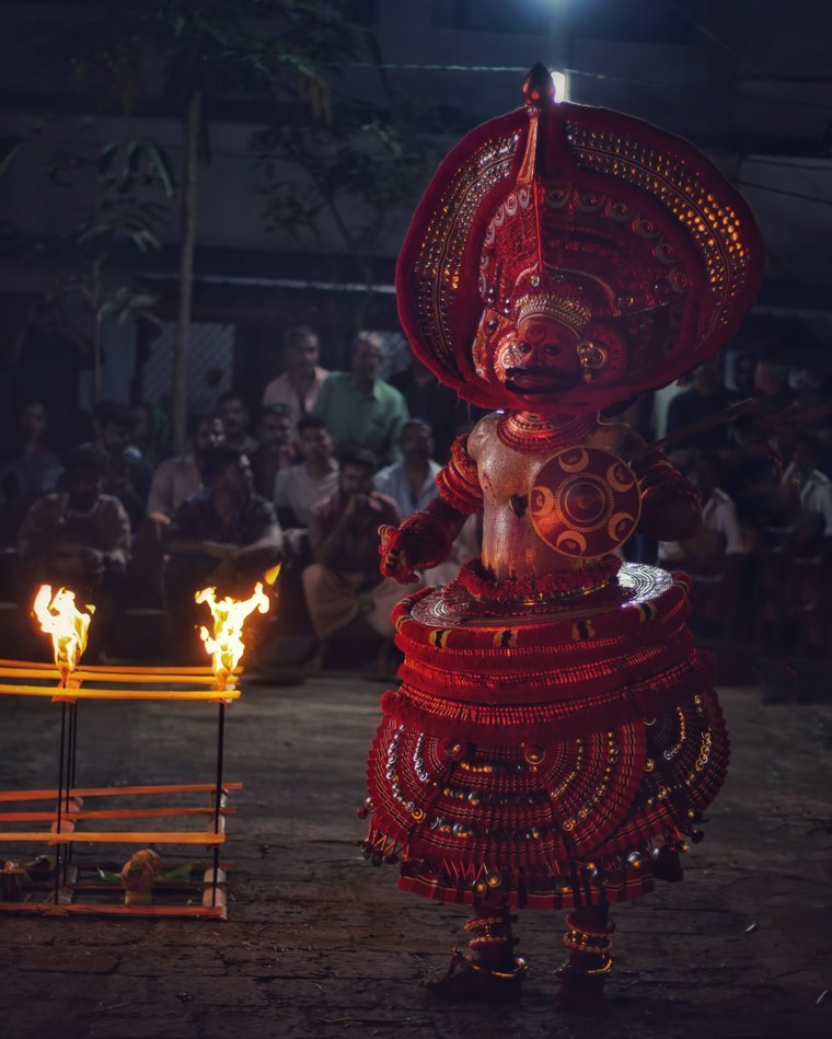 Kudiveeran Theyyam at night in a red halo-shaped headdress and layered red attire, with a lamp stand in front