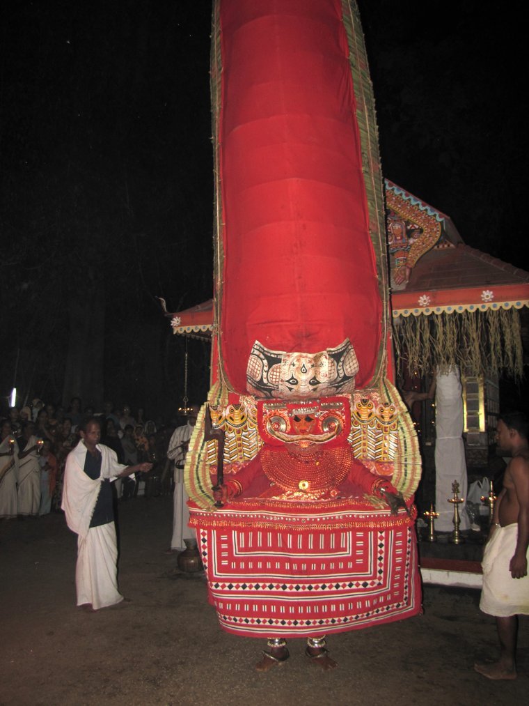Thai Paradevatha Theyyam at night with a very tall red crown, at a shrine in Kannur