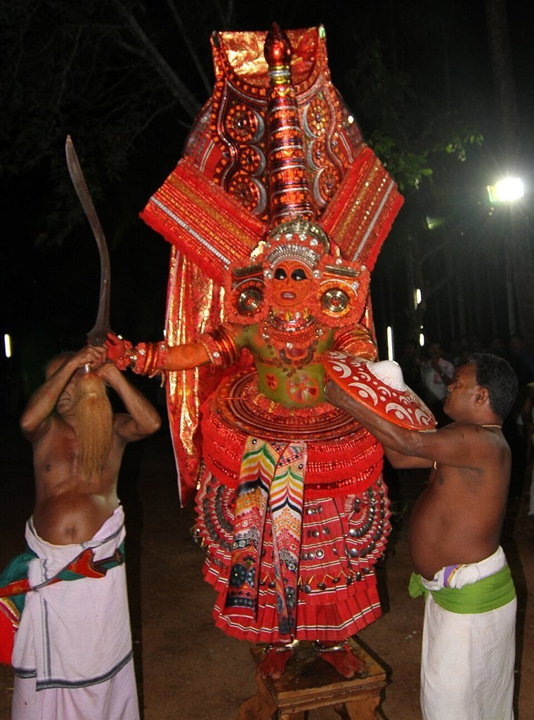 Vairajathan Theyyam in a tall red crown, holding a sword and shield, with attendants beside him
